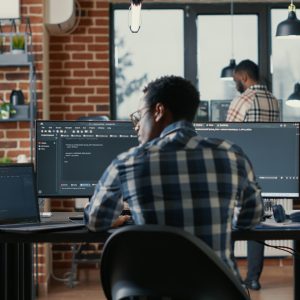 Programer coding on laptop sitting down at desk with computer screens parsing code in software agency. Software developer compiling algorithms with cloud programers working in the background.